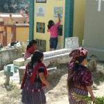 Local women head into the Chichicastenango General Cemetery with flowers to place on graves in preparation for the Dia De Los Muertos. In the background other women paint a tomb.
