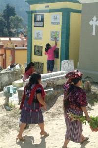 Local women head into the Chichicastenango General Cemetery with flowers to place on graves in preparation for the Dia De Los Muertos. In the background other women paint a tomb.